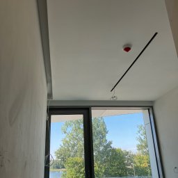 Usługi wykończeniowe- Robert Nadolski - Surowa ściana z widocznymi śladami szpachlowania, z oknem na jezioro. Na suficie czujnik dymu i czarna listwa oświetleniowa z wystającymi kablami. Wnętrze w trakcie wykańczania.