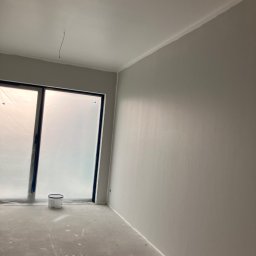 Usługi wykończeniowe- Robert Nadolski - Pomieszczenie po malowaniu na szaro. Okno zabezpieczone folią, widoczne ślady farby na podłodze i puszka farby. Minimalistyczne wykończenie wnętrza.