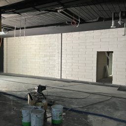 Usługi wykończeniowe- Robert Nadolski - Wnętrze w trakcie malowania: ściana z cegieł pomalowana na biało, widoczne puszki z farbą Teknos Ekora i sprzęt do malowania natryskowego. Sufit w kolorze czarnym.