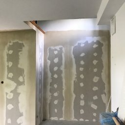 Usługi wykończeniowe- Robert Nadolski - Wnętrze w trakcie prac: ściany częściowo pokryte warstwą szpachli, widoczne miejsca na gniazdka elektryczne, surowy beton na podłodze i materiały budowlane w rogu.