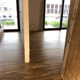 Usługi wykończeniowe- Robert Nadolski - Jasne drewniane panele podłogowe w nowym wnętrzu z widokiem na zewnątrz przez duże okna. Panele Artens opierają się o ścianę.