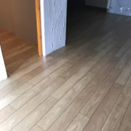 Jasne panele podłogowe z widoczną strukturą drewna, ułożone wzdłuż ściany z dekoracyjną, białą fakturą. W tle grzejnik i fragment okna.