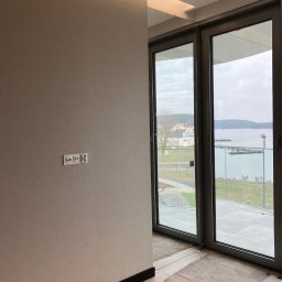 Usługi wykończeniowe- Robert Nadolski - Pokój po remoncie z widokiem na jezioro przez duże okna. Ściana w beżowym odcieniu, widoczne gniazdka elektryczne. Podłoga zabezpieczona kartonem.