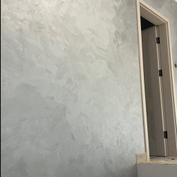 Usługi wykończeniowe- Robert Nadolski - Ściana z efektem dekoracyjnym w odcieniach szarości, widoczna obok otwartych drzwi. Wykończenie wnętrza z charakterystyczną fakturą.