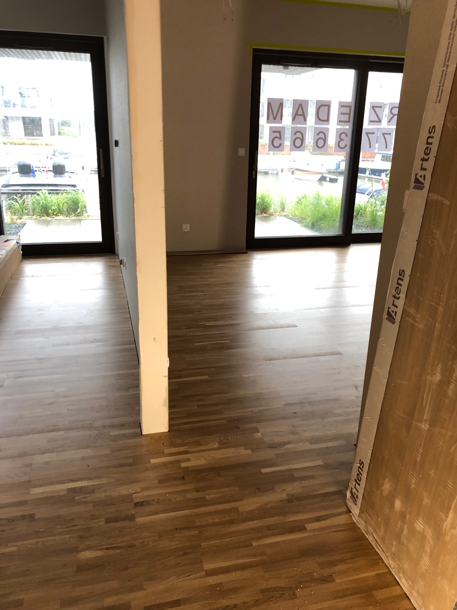 Jasne drewniane panele podłogowe w nowym wnętrzu z widokiem na zewnątrz przez duże okna. Panele Artens opierają się o ścianę.