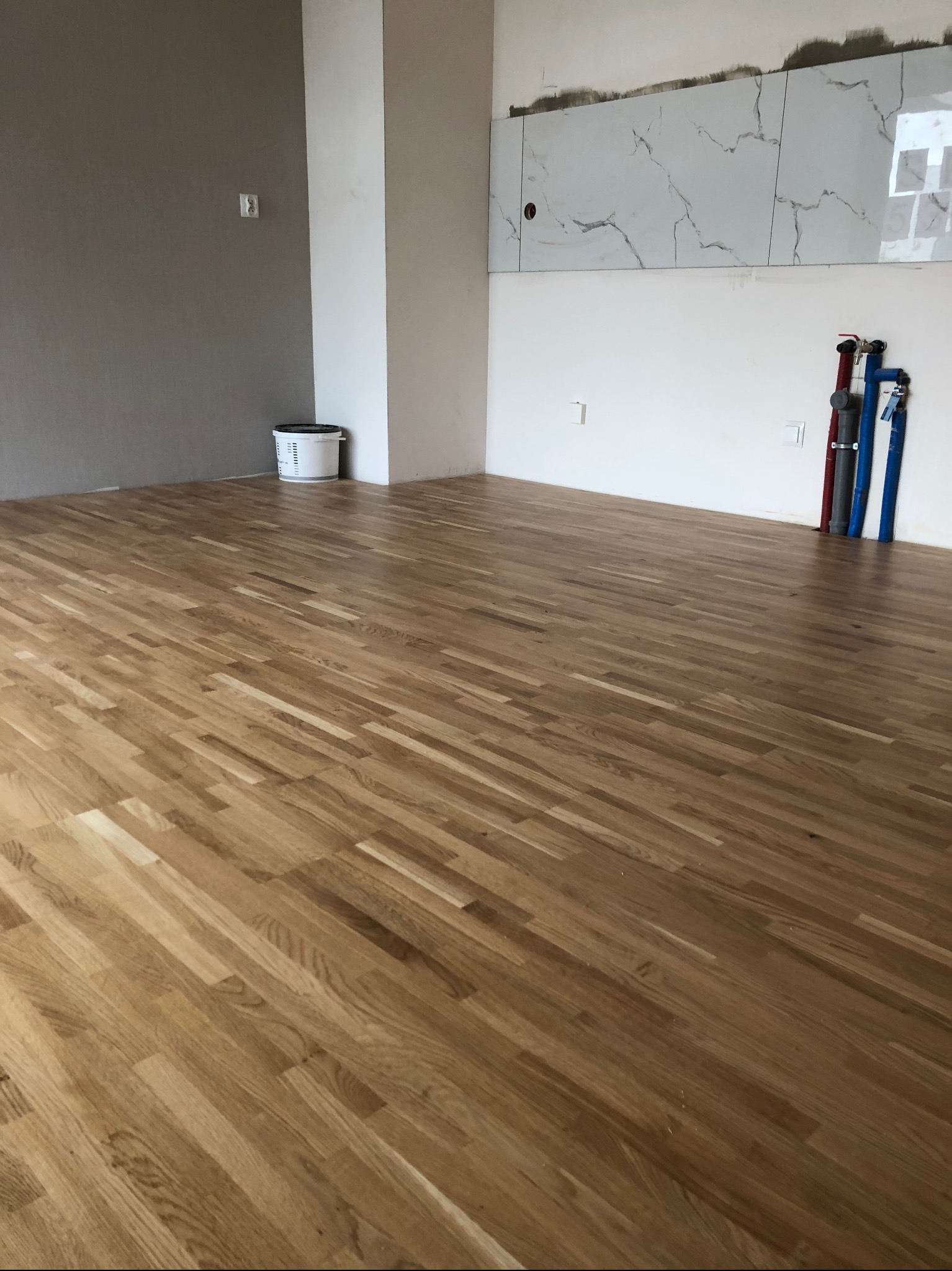 Jasne panele ułożone w przestronnym pokoju z surowymi ścianami i fragmentem marmurowej mozaiki. Widoczne piony instalacji wodnej i wiadro farby w rogu.