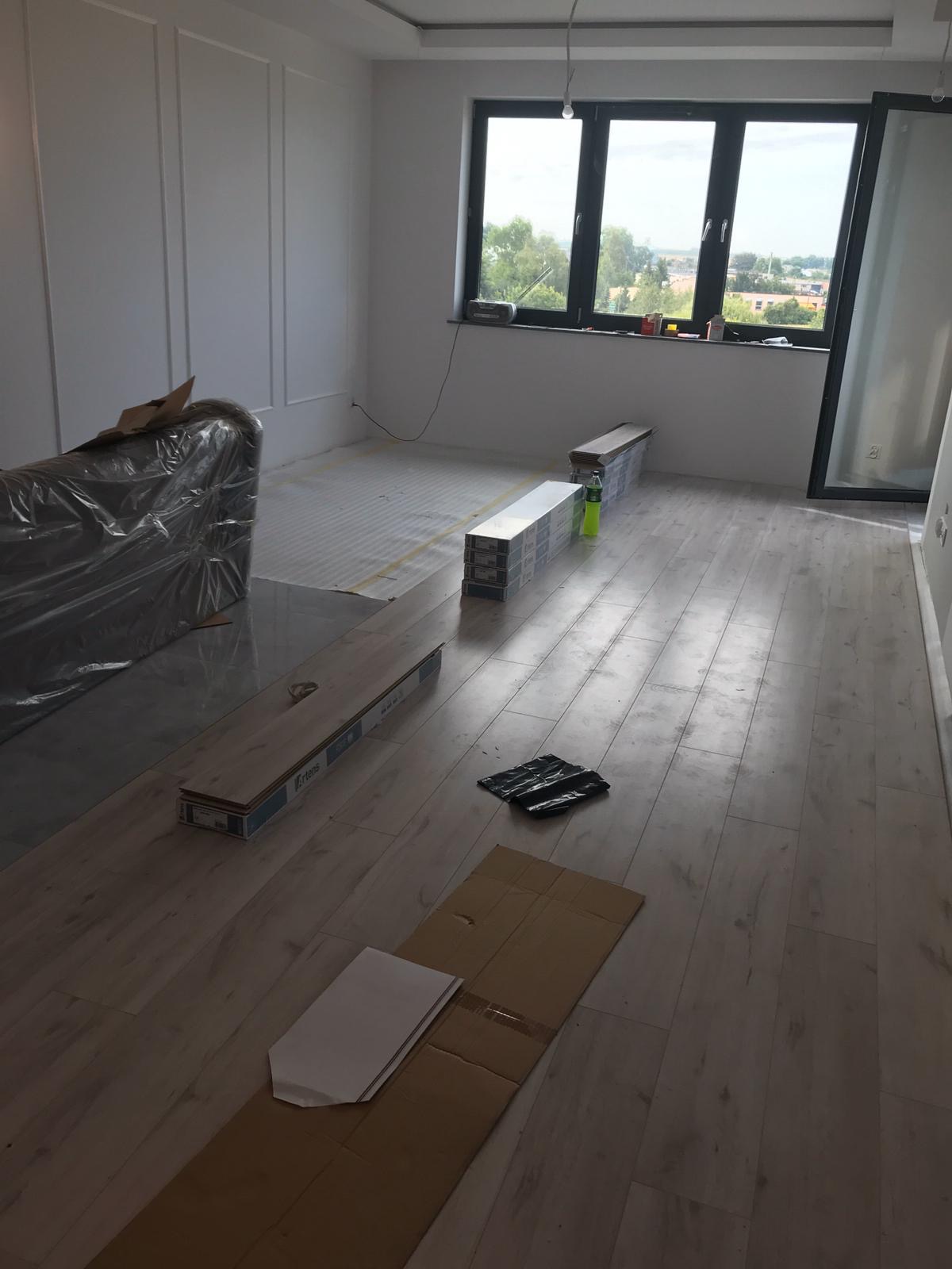 Jasne panele podłogowe w trakcie montażu w przestronnym pokoju z dużym oknem. Część podłogi wyłożona, widoczne paczki paneli i narzędzia.