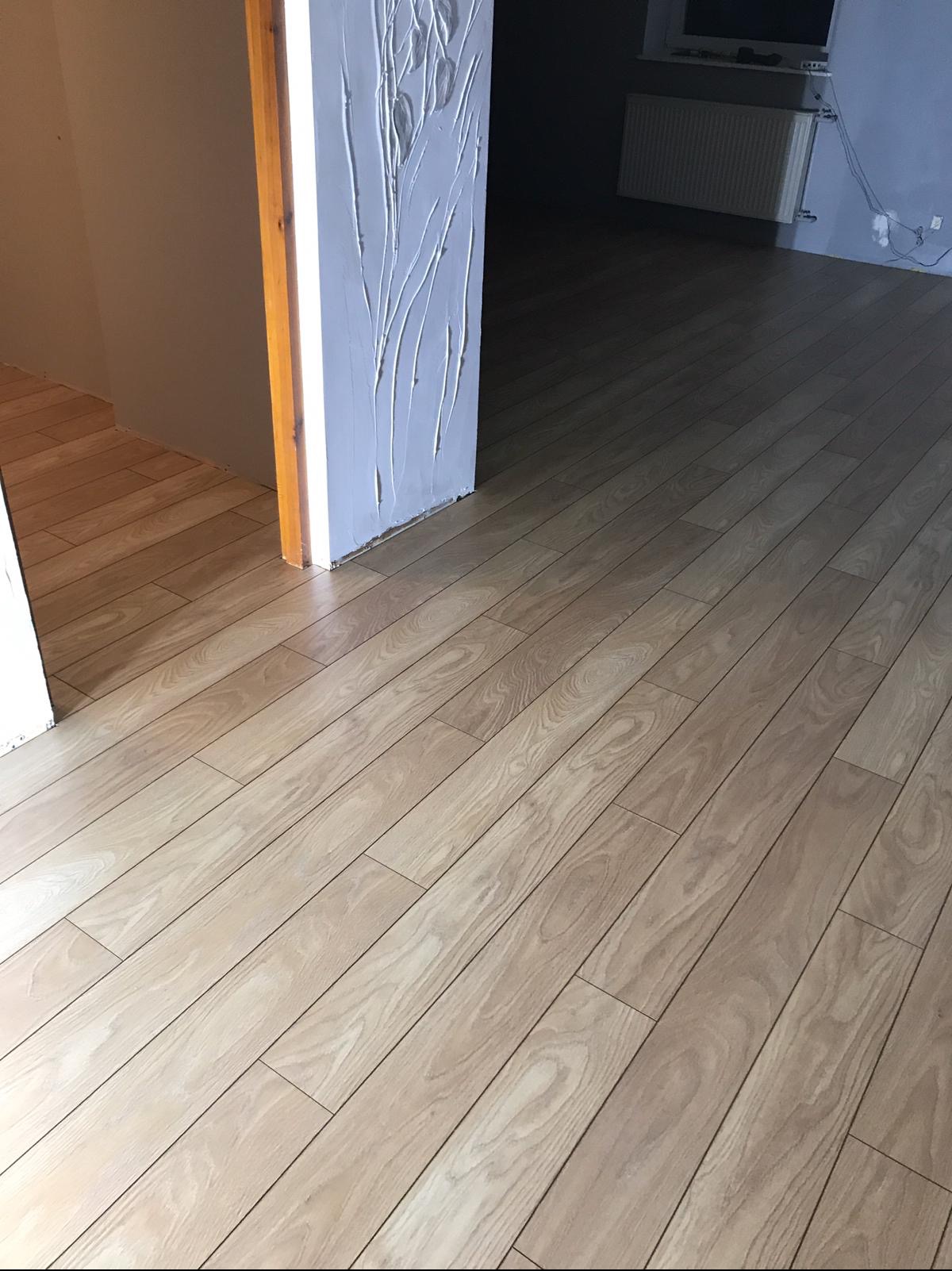 Jasne panele podłogowe z widoczną strukturą drewna, ułożone wzdłuż ściany z dekoracyjną, białą fakturą. W tle grzejnik i fragment okna.