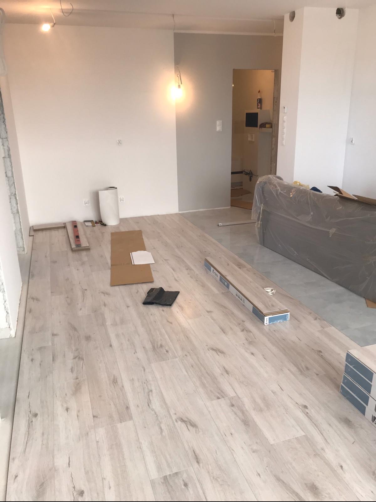 Jasne panele podłogowe w trakcie układania. Widoczne narzędzia, kartony z panelami i częściowo zakryty mebel. Ściany białe, surowe oświetlenie.