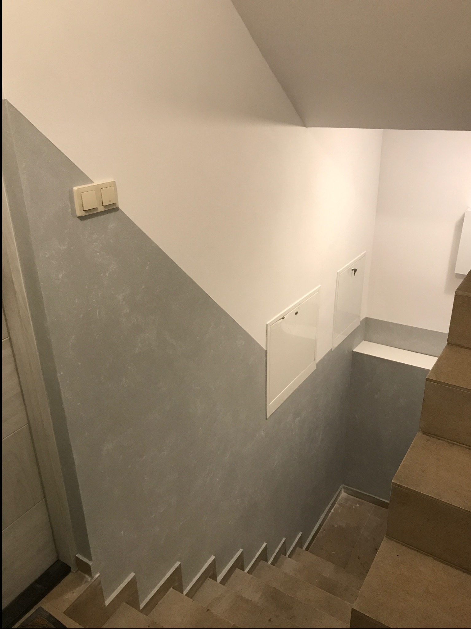 Klatka schodowa po remoncie: ściany pomalowane w dwóch odcieniach szarości, z widocznymi schodami i drzwiczkami rewizyjnymi. Nowoczesne wykończenie.