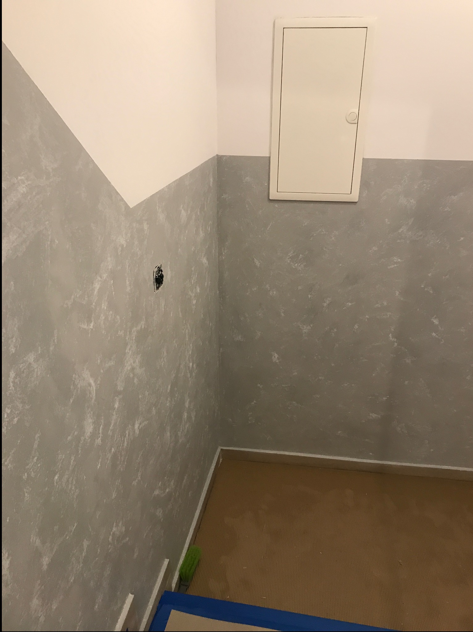 Wykończony narożnik pokoju z dekoracyjną szarą ścianą, białą górą i brązową podłogą. Widoczna klapka rewizyjna i gniazdko elektryczne. Częściowo widoczna szczotka i taśma malarska.