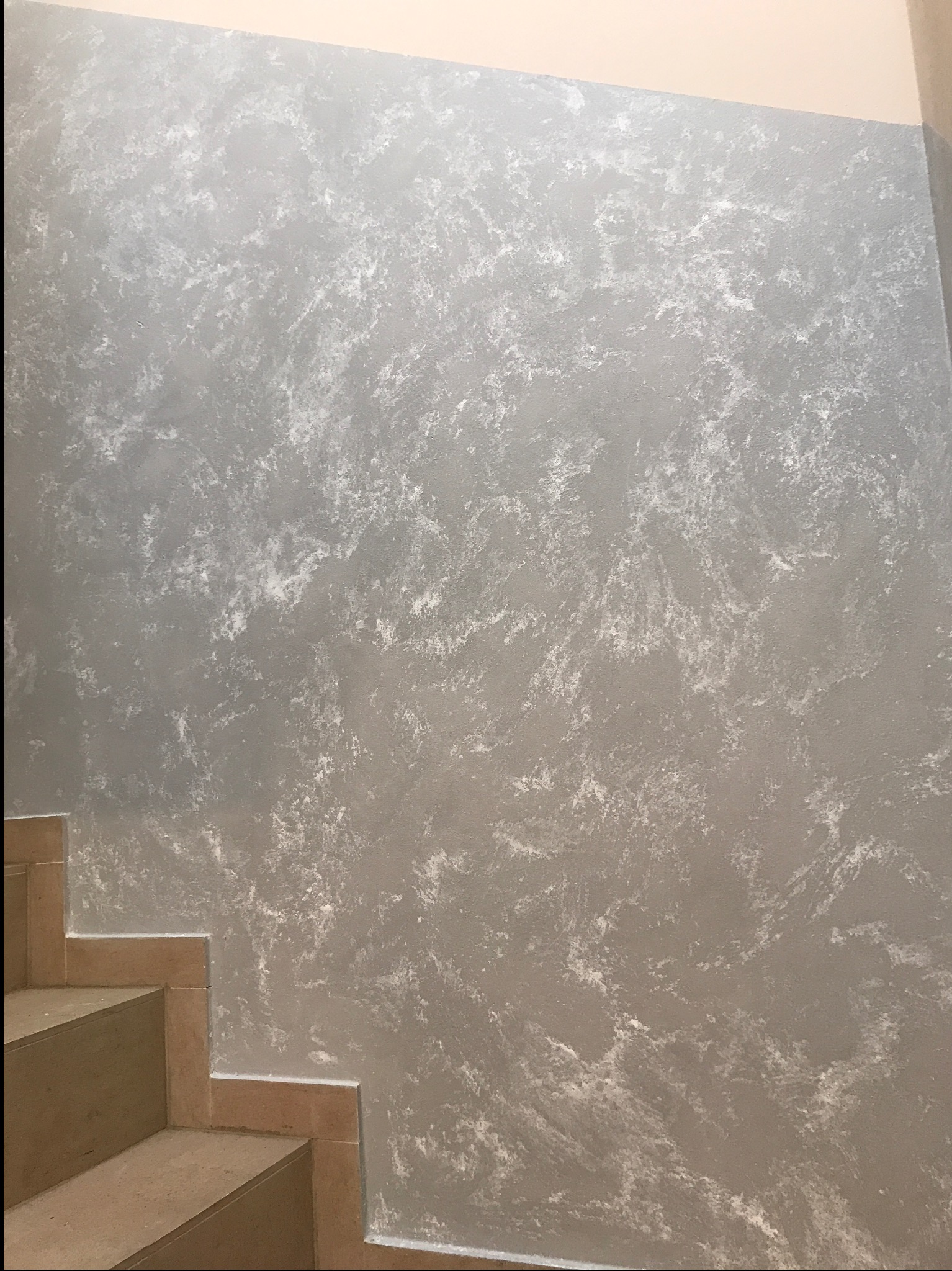 Ściana w odcieniach szarości z dekoracyjną fakturą, widoczna obok fragmentu schodów z beżowymi stopniami. Efektowna aranżacja wnętrza.