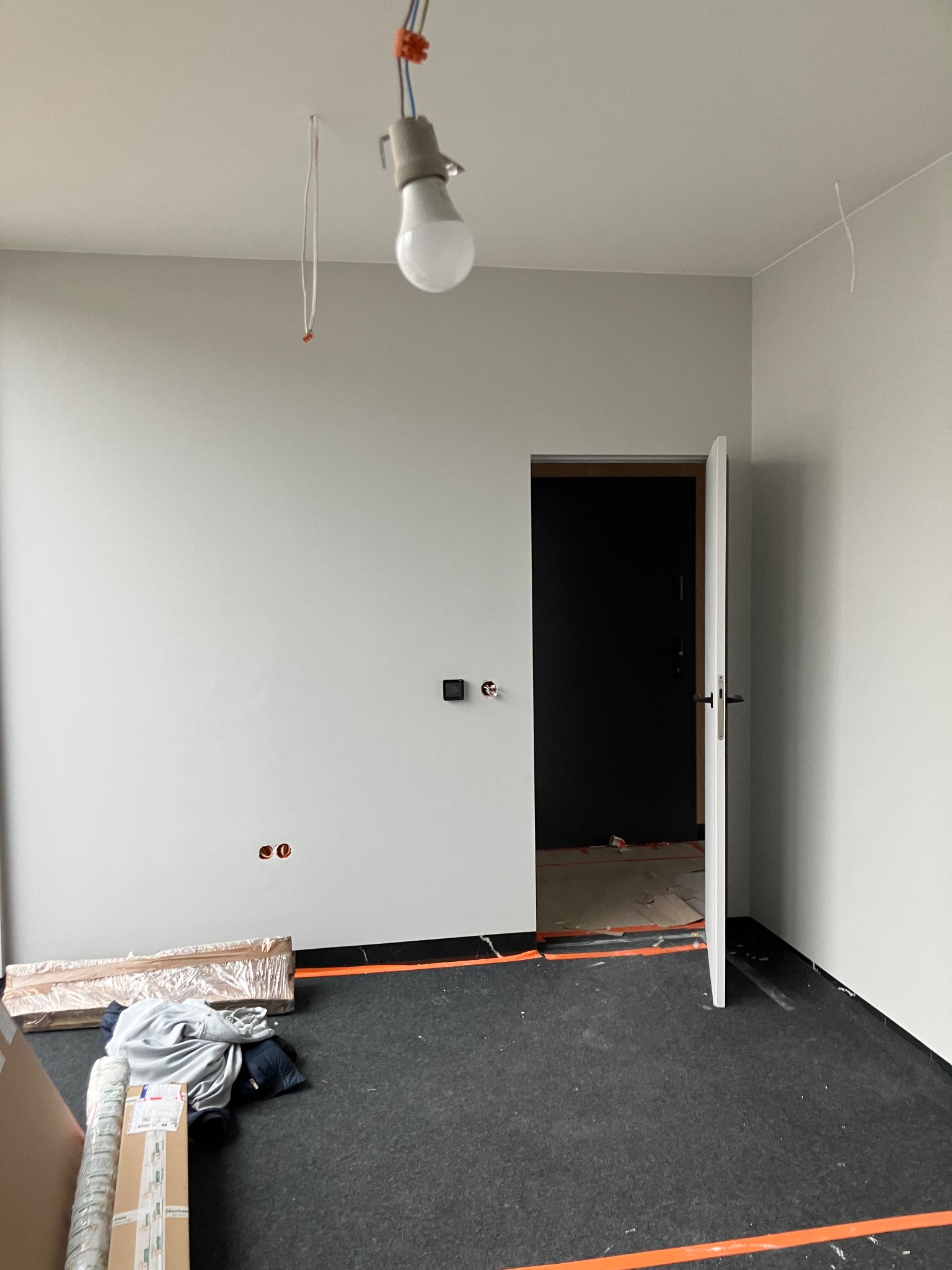 Wnętrze pokoju w trakcie remontu: gładkie, szare ściany, czarna podłoga z widocznymi przewodami, otwarte białe drzwi i wisząca żarówka. Widoczne materiały budowlane.