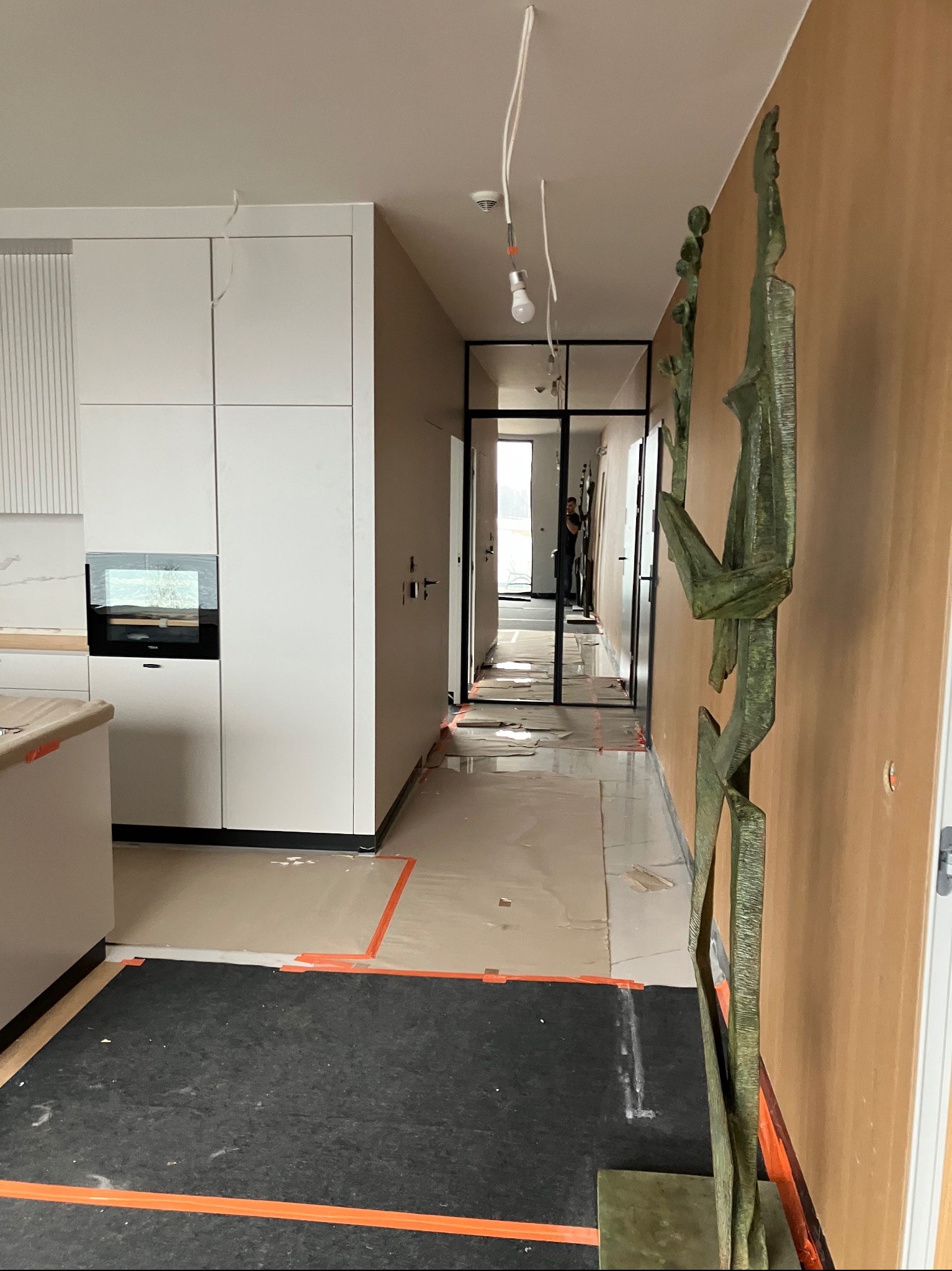 Wnętrze w trakcie prac wykończeniowych: zabezpieczona podłoga, nowoczesna kuchnia z białymi szafkami, metalowa rzeźba przy ścianie. Widoczne przygotowania do dalszych prac.