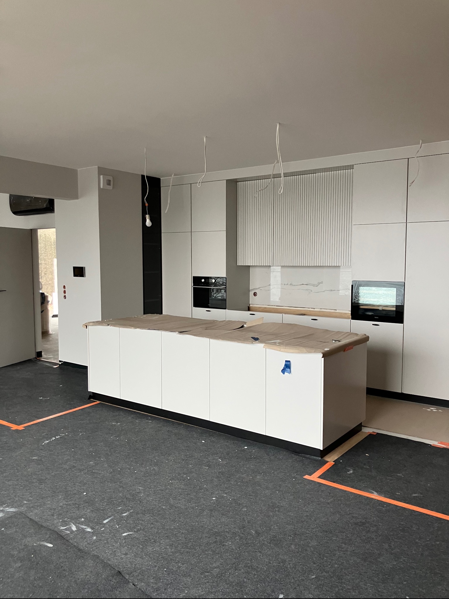 Nowoczesna, jasna kuchnia w trakcie montażu: białe szafki, wyspa kuchenna zabezpieczona folią, widoczne instalacje elektryczne i fragmenty podłogi z zabezpieczeniami.