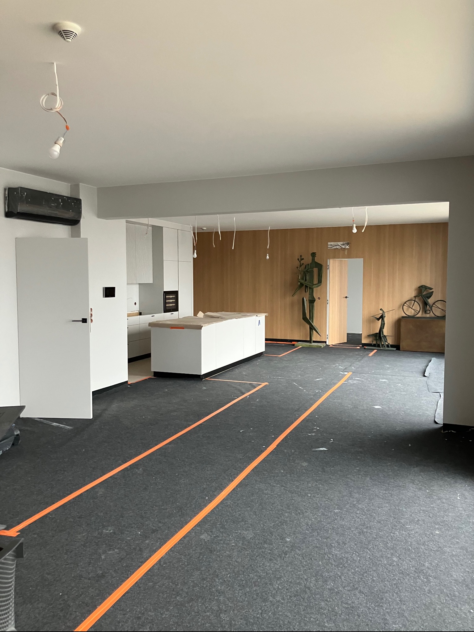 Wnętrze w trakcie prac wykończeniowych: zabezpieczona podłoga, widoczna kuchnia z wyspą, drewniana ściana z rzeźbami. Wiszące kable oświetleniowe.