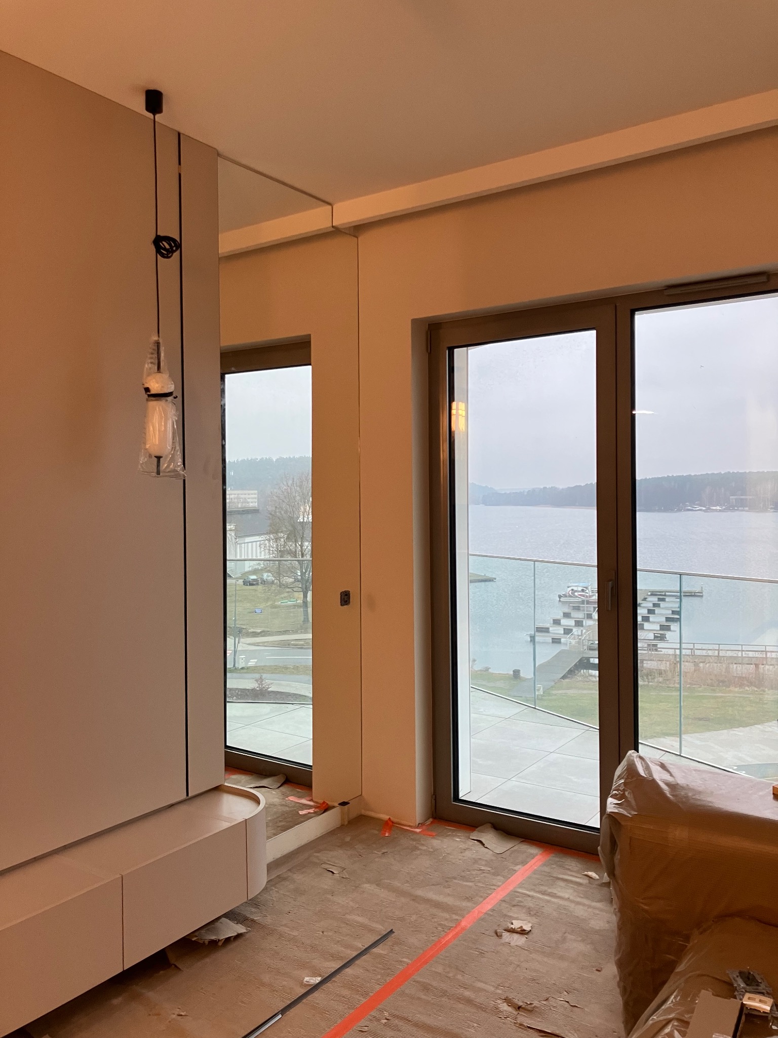 Wnętrze w trakcie prac wykończeniowych: widok na jezioro przez okno, podłoga przygotowana pod panele, minimalistyczna zabudowa meblowa, lampa wisząca w folii ochronnej.
