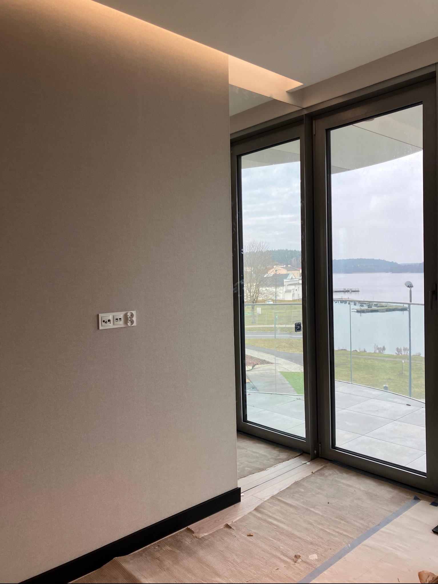 Pokój po remoncie z widokiem na jezioro przez duże okna. Ściana w beżowym odcieniu, widoczne gniazdka elektryczne. Podłoga zabezpieczona kartonem.