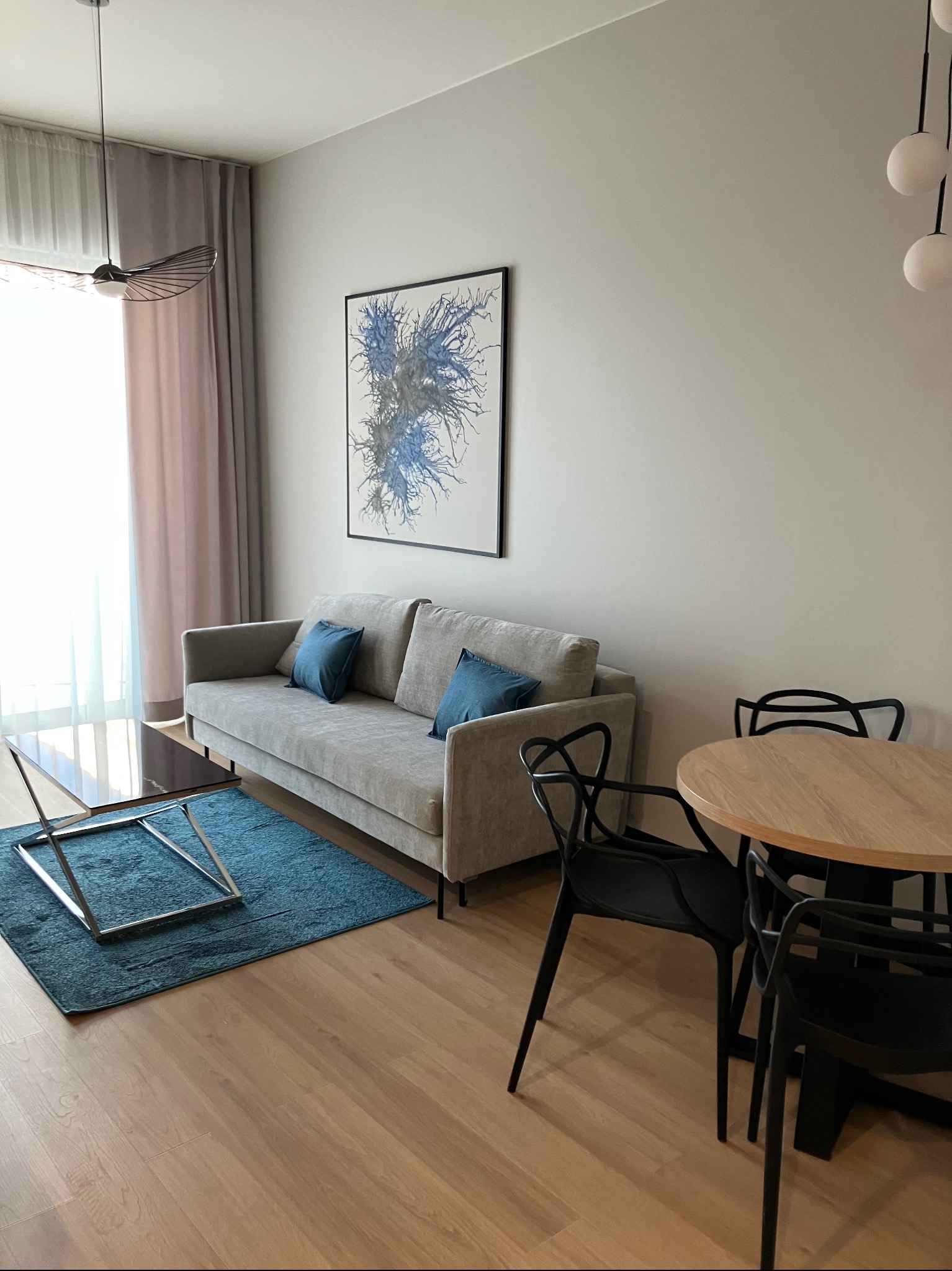 Jasne wnętrze salonu z drewnianą podłogą, szarą sofą z niebieskimi poduszkami, okrągłym stołem i czarnymi krzesłami. Na ścianie abstrakcyjny obraz w czarnej ramie.