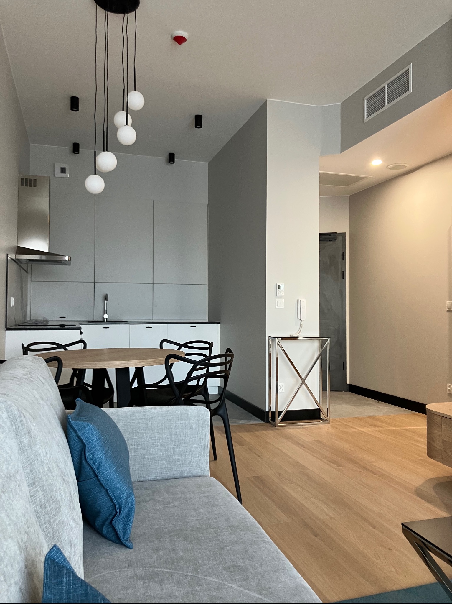 Nowoczesne wnętrze mieszkania z drewnianą podłogą, szarą zabudową kuchenną i okrągłym stołem. Minimalistyczny design z wiszącymi lampami kulami.