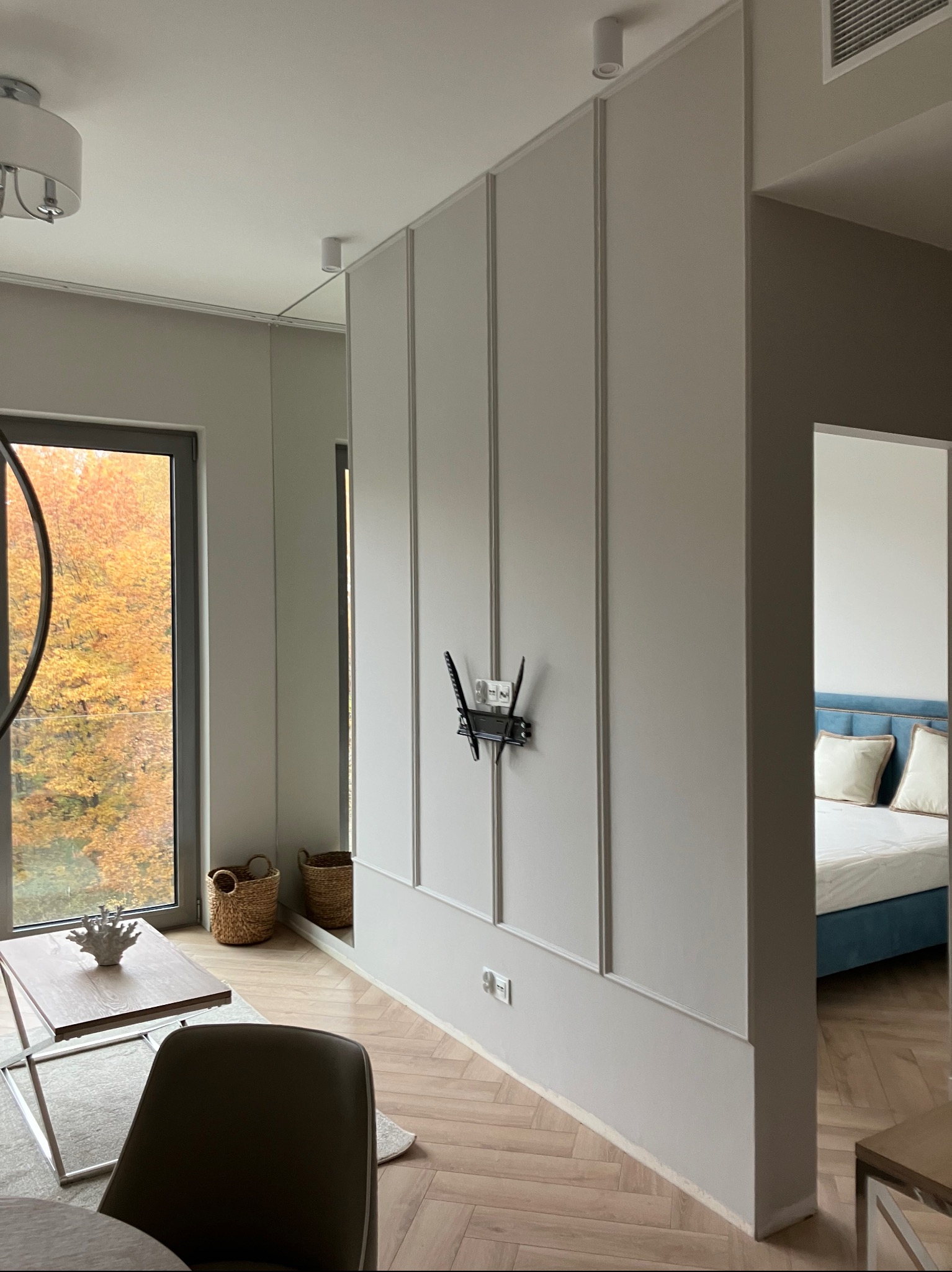 Eleganckie wnętrze: szara ściana z dekoracyjnymi panelami, uchwyt TV, parkiet w jodełkę, widok na salon i jesienny krajobraz za oknem. Minimalistyczny design.