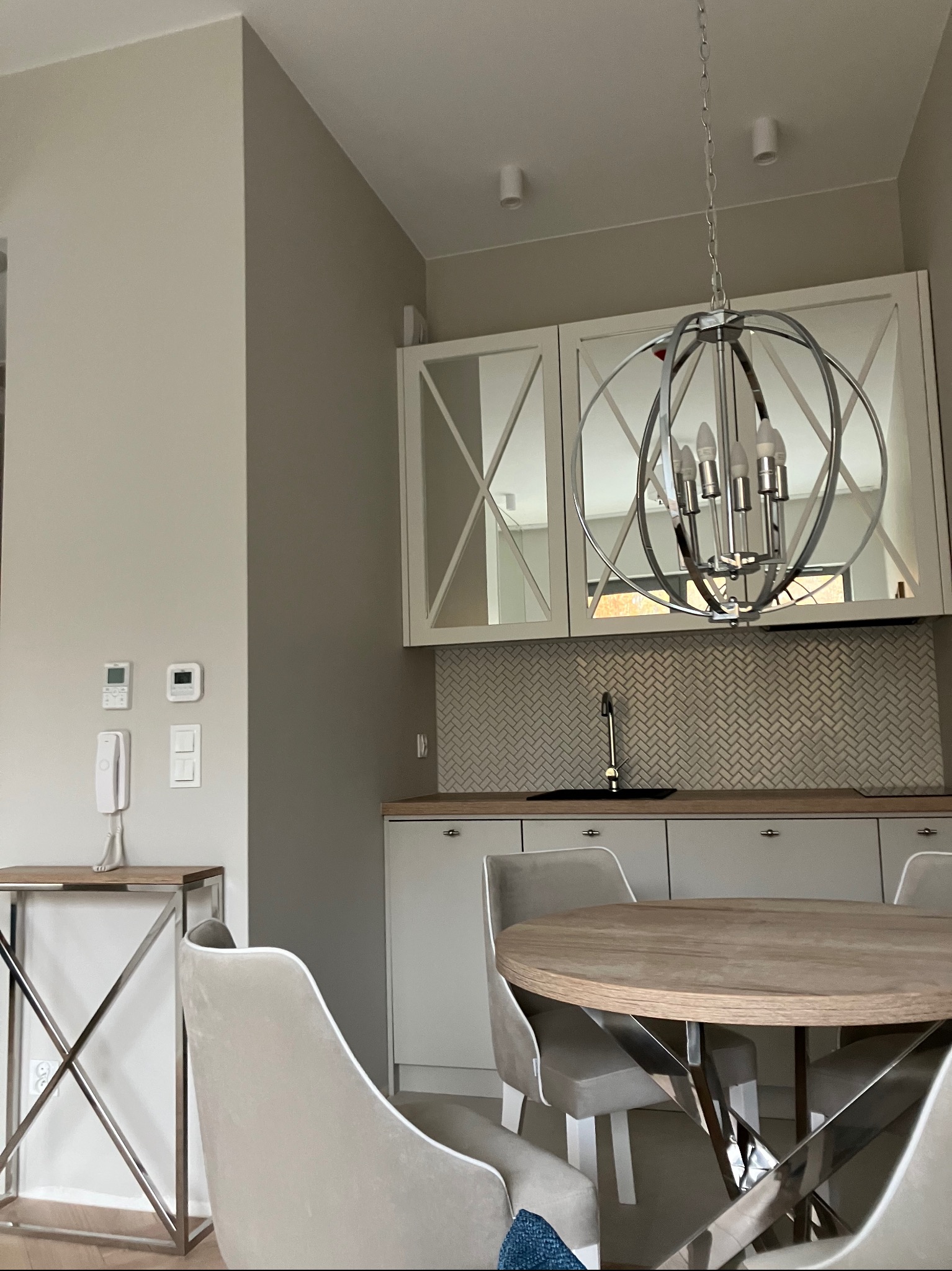 Jasne wnętrze kuchni z okrągłym stołem i krzesłami. Nad stołem wisi nowoczesna lampa. Na ścianie białe szafki z lustrzanymi frontami i mozaika w jodełkę.