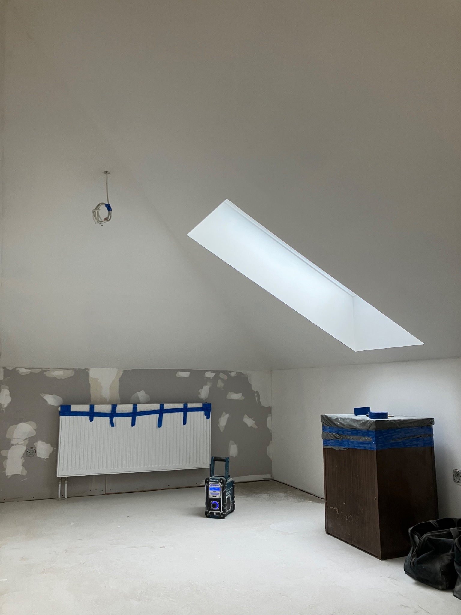 Pokój na poddaszu w trakcie remontu: zabezpieczony grzejnik taśmą, ściany częściowo wyszpachlowane, okno dachowe, radio budowlane i torba narzędziowa.