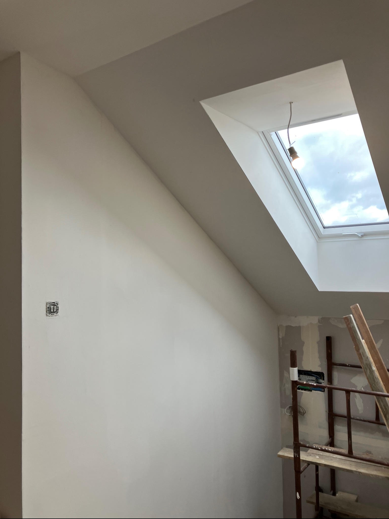 Ściana po remoncie z oknem dachowym i widoczną instalacją elektryczną. Fragment rusztowania w rogu kadru. Świeżo pomalowana powierzchnia.