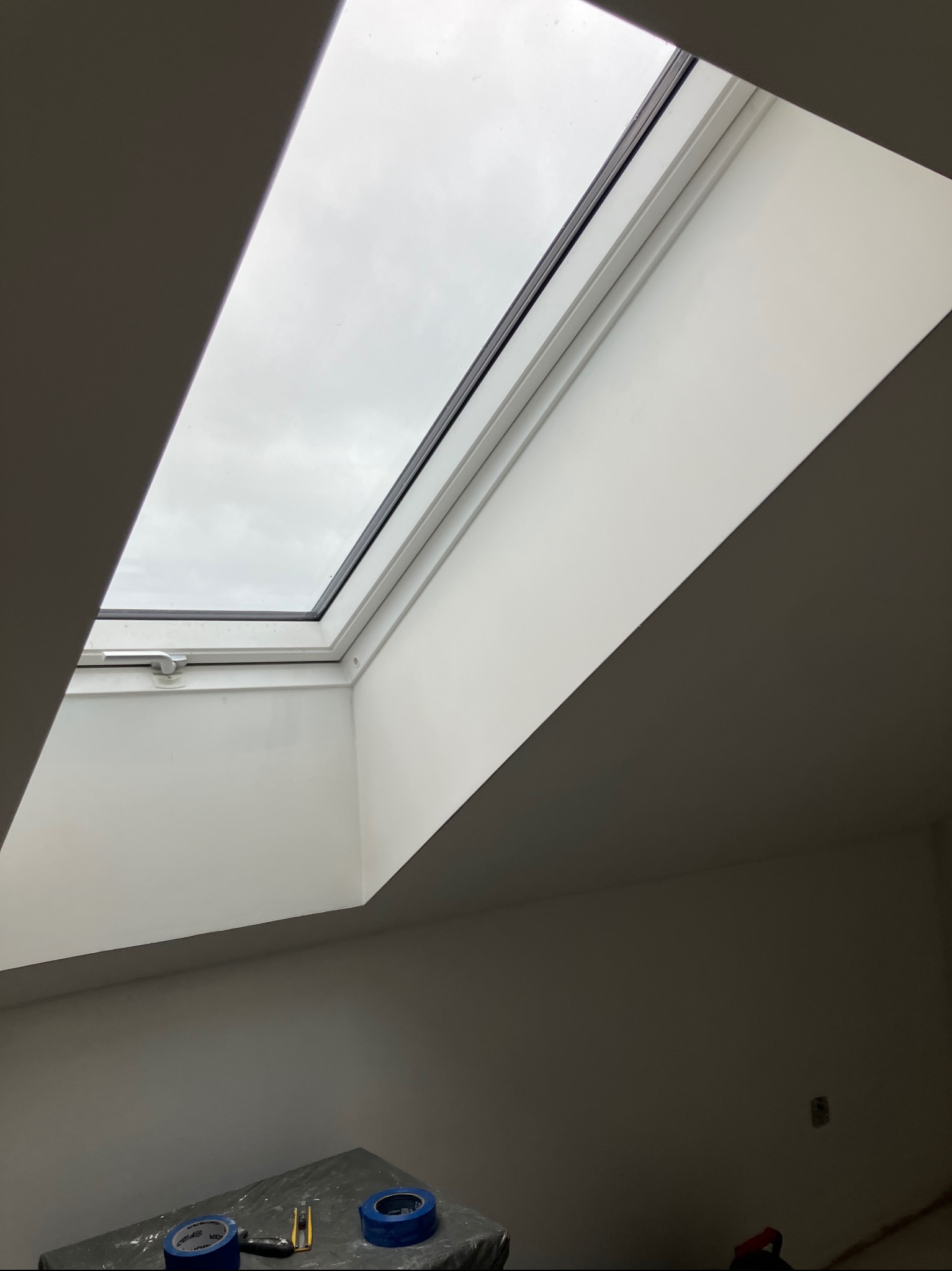 Białe ściany po malowaniu wokół okna dachowego. Na dole widoczne narzędzia malarskie: taśmy malarskie i nożyk na zabezpieczonej powierzchni.