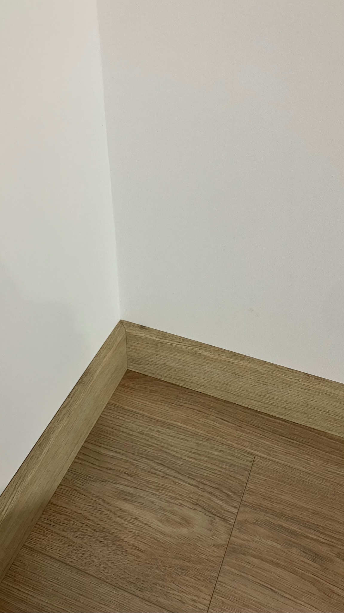 Jasne panele podłogowe z listwami przypodłogowymi w rogu pomieszczenia z białymi ścianami. Widok z góry, neutralne oświetlenie.