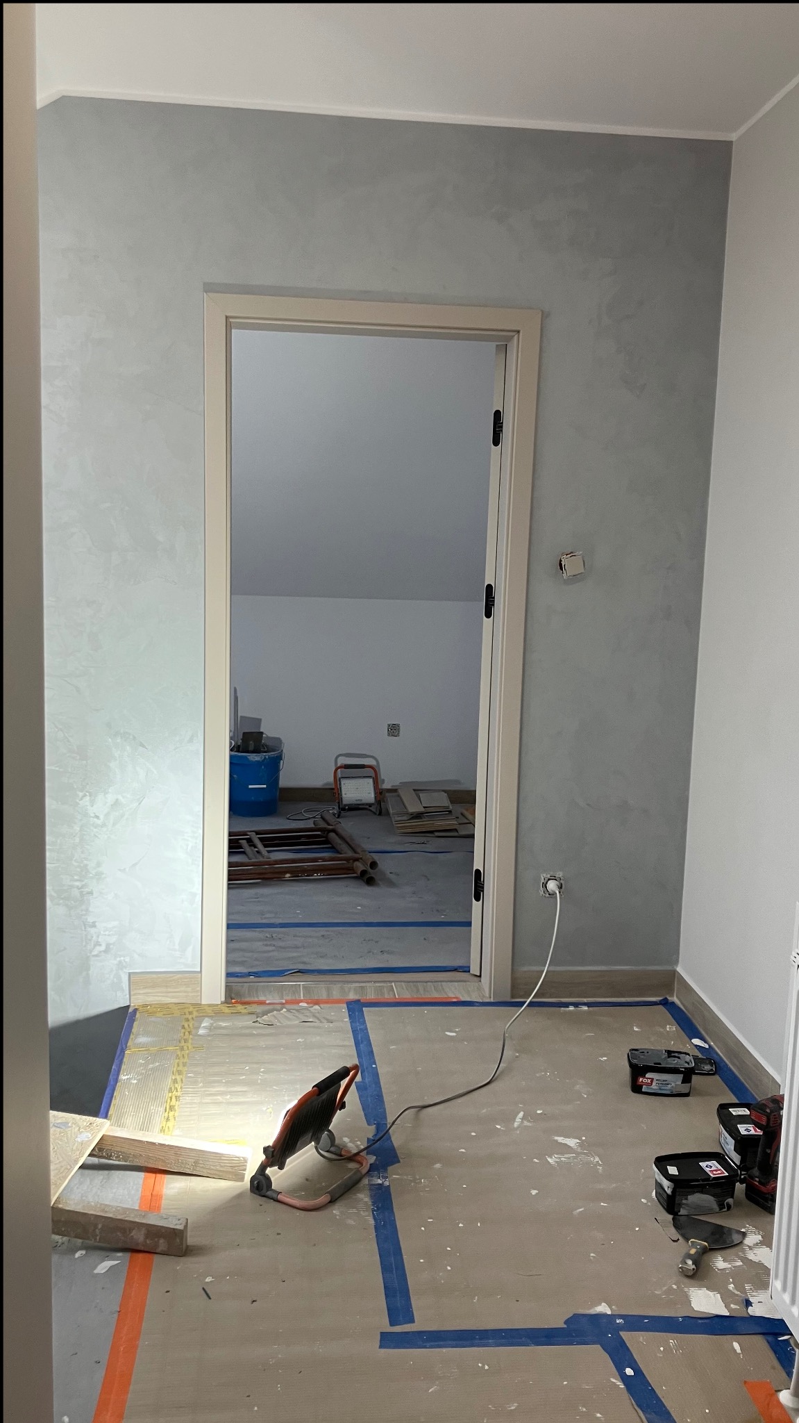 Pokój w trakcie remontu: świeżo pomalowane ściany w odcieniach szarości, zabezpieczona podłoga, widoczne narzędzia i materiały budowlane, otwarte drzwi do kolejnego pomieszczenia.