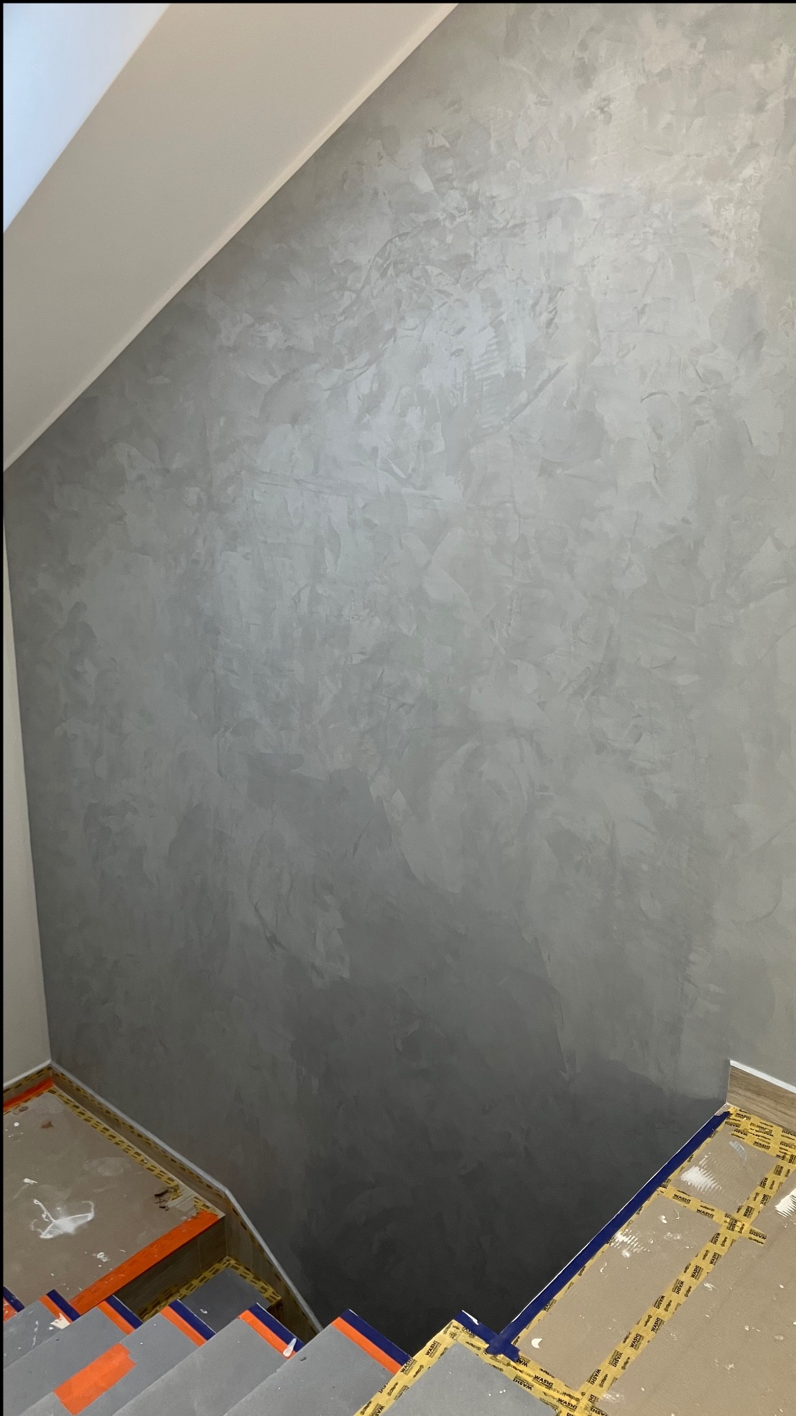Ściana w odcieniach szarości z dekoracyjną fakturą, widoczna z perspektywy schodów zabezpieczonych taśmą malarską. Minimalistyczny design wnętrza.
