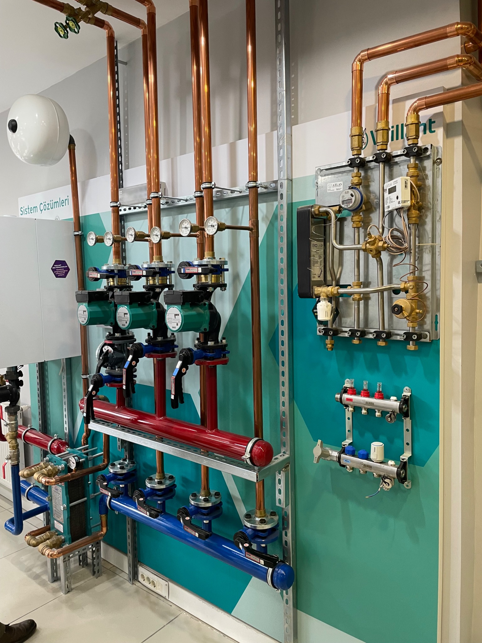 Złożona instalacja grzewcza z miedzianymi rurami, zielonymi pompami i manometrami na tle ściany w odcieniach turkusu i bieli. Widoczne liczniki i zawory.