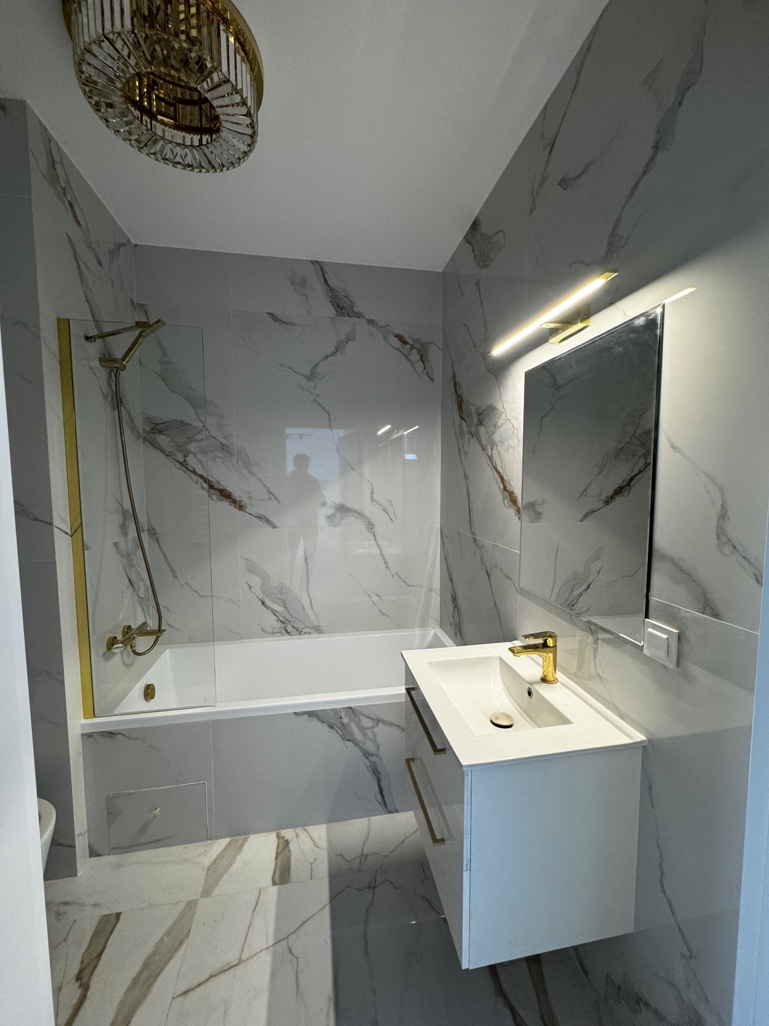 Biała łazienka z marmurowymi płytkami, złotą armaturą i oświetleniem. Nowoczesna umywalka z szafką, wanna z parawanem i ozdobny żyrandol. Eleganckie wykończenie wnętrza.