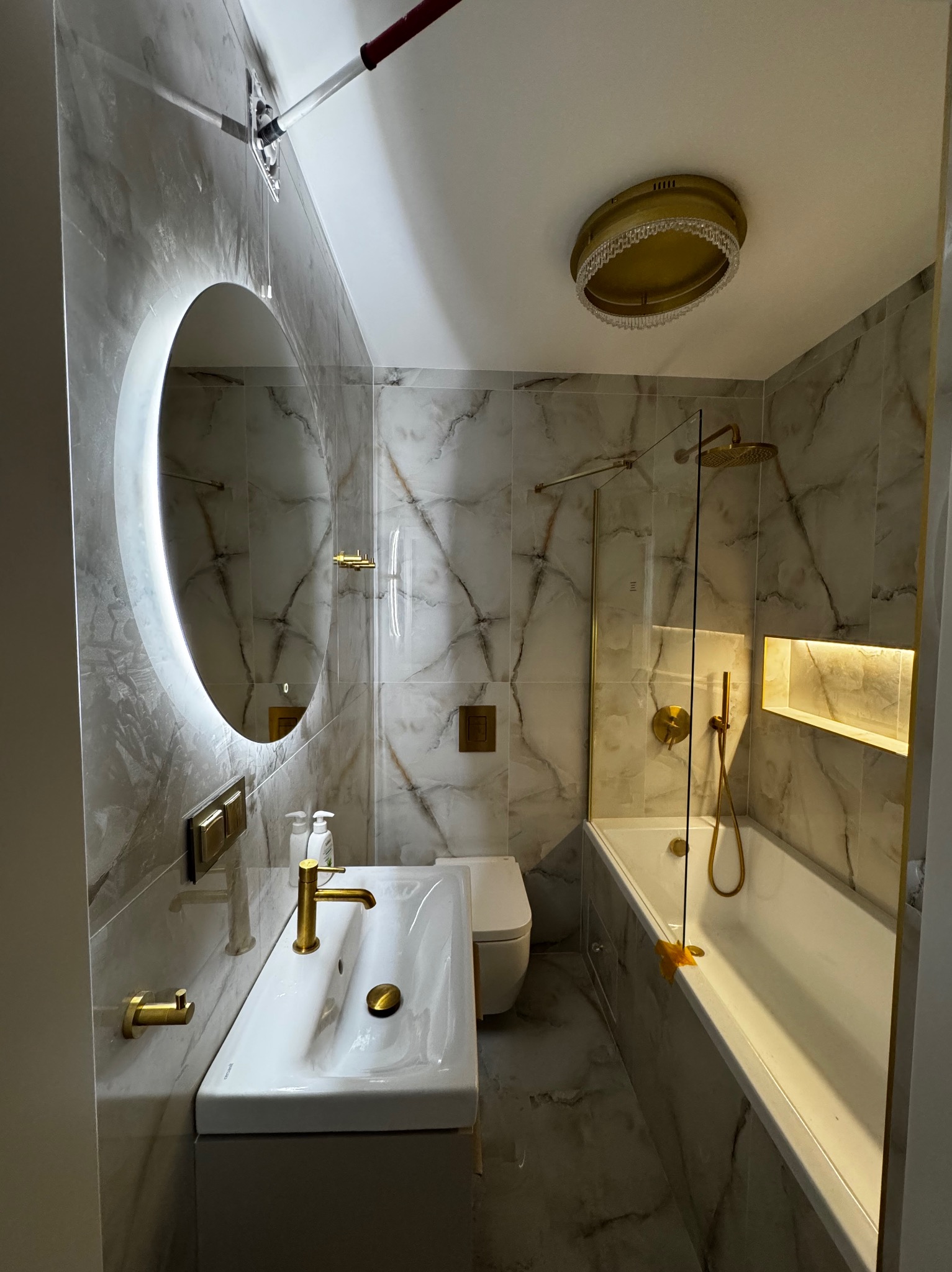 Elegancka łazienka w szarościach z marmurowym wzorem, złote akcenty armatury i oświetlenia, okrągłe lustro z podświetleniem LED, nowoczesny design.