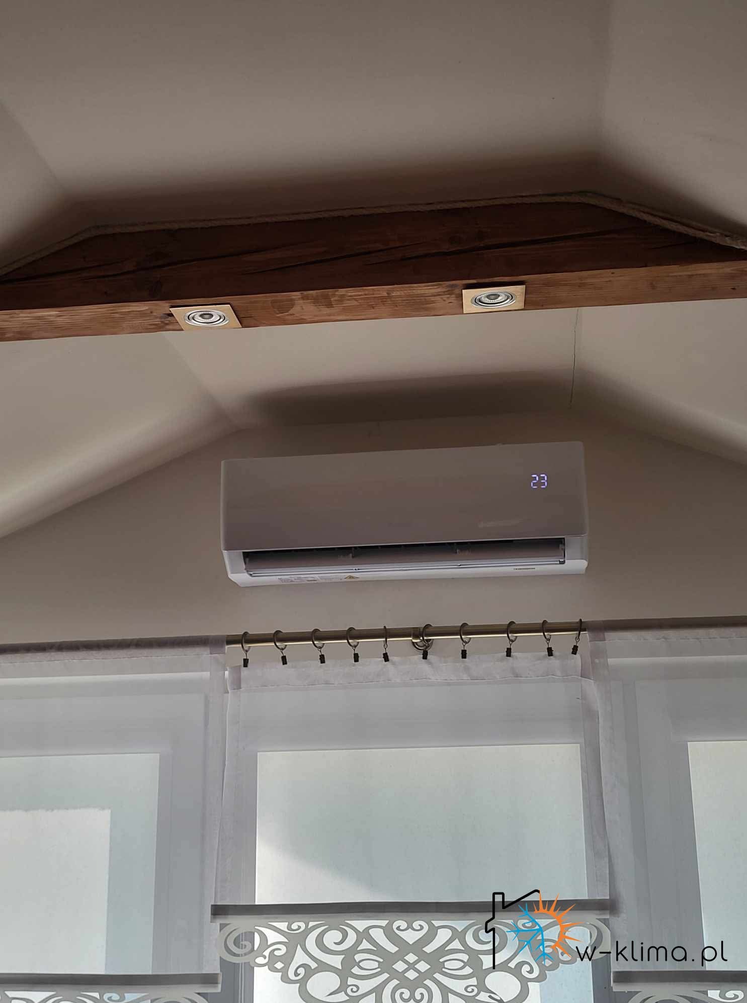 Klimatyzator ścienny z wyświetlaczem temperatury (23°C) zamontowany w jasnym pomieszczeniu z drewnianymi belkami stropowymi i zasłonami.