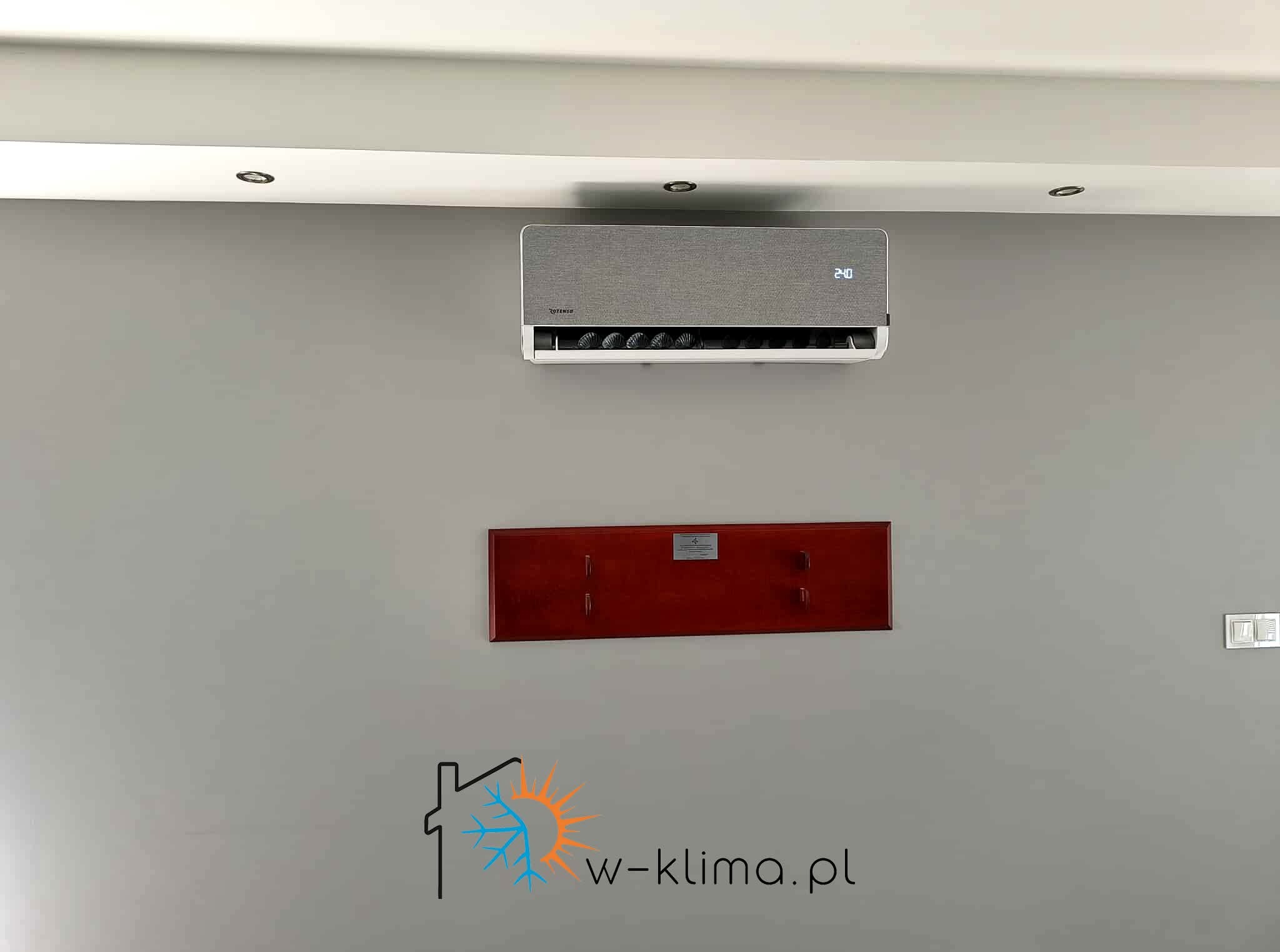Szary klimatyzator Cotensu na ścianie w pokoju z oświetleniem punktowym i wieszakiem. Na ścianie widoczne logo firmy w-klima.pl.