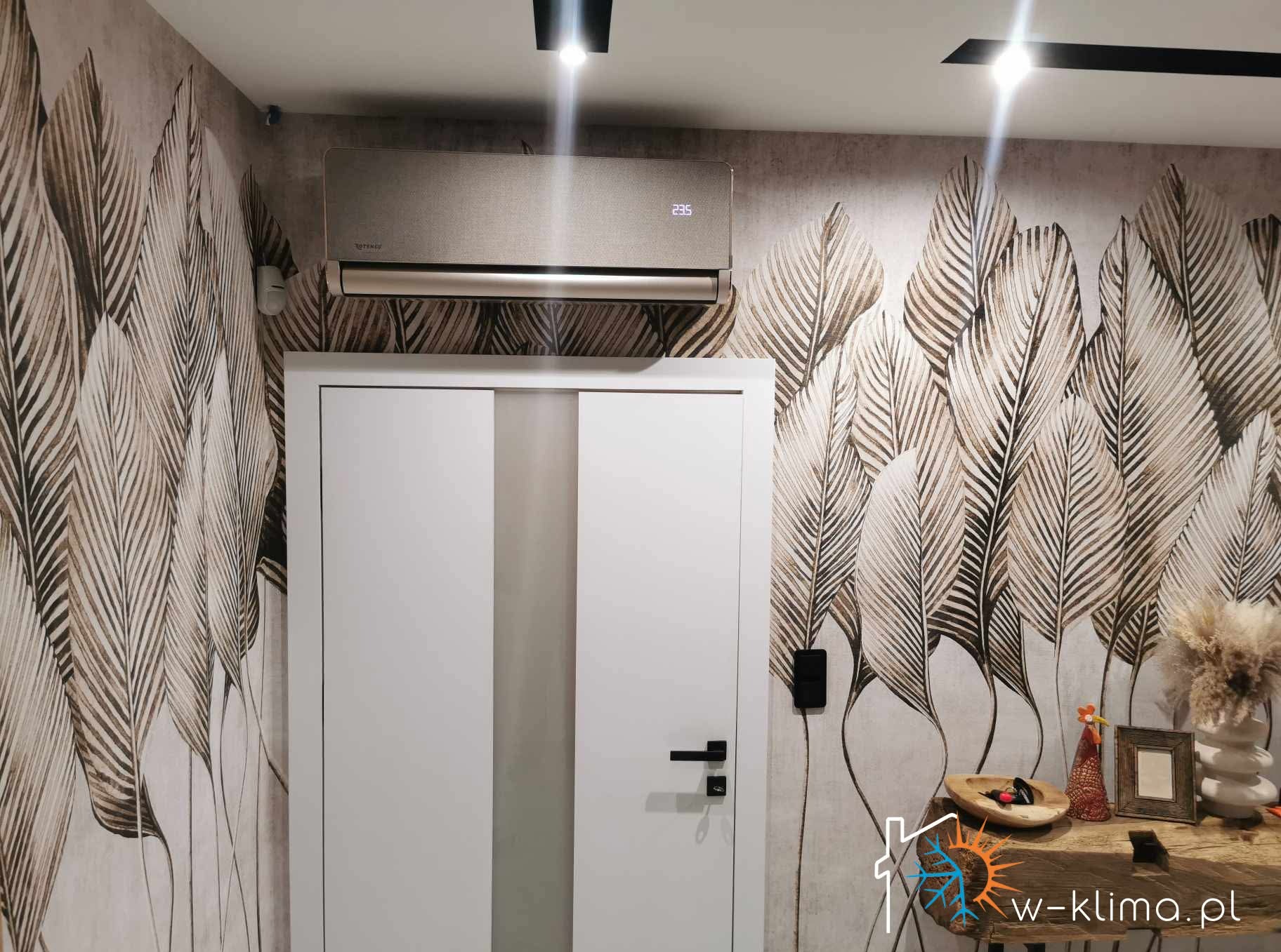Klimatyzator w kolorze szampańskim zamontowany nad białymi drzwiami w pomieszczeniu z tapetą w liście. Widoczny fragment stołu z dekoracjami. Nowoczesny design, ciepłe oświetlenie.
