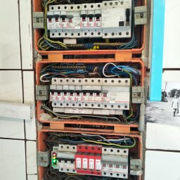 Przemysław Kozłowski - Rozdzielnia elektryczna w pomarańczowej obudowie, widoczne bezpieczniki, przewody i listwy. Instalacja zamontowana w ścianie z białymi płytkami. Widoczne elementy instalacji elektrycznej.