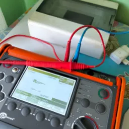 Miernik Sonel MPI-530-IT z podłączonymi przewodami pomiarowymi, wyświetlacz pokazuje wynik pomiaru rezystancji izolacji. Obok skrzynka elektryczna i ładowarka.