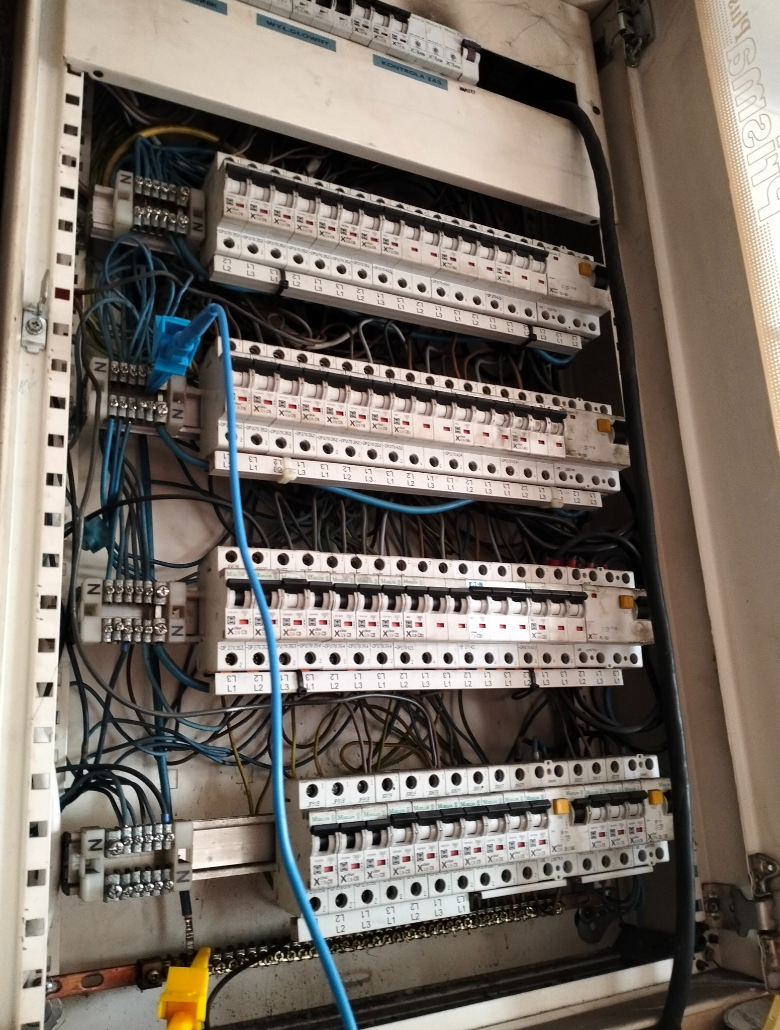 Rozdzielnica elektryczna z widocznymi bezpiecznikami, przewodami i listwami zaciskowymi wewnątrz obudowy. Widoczne oznaczenia na elementach.