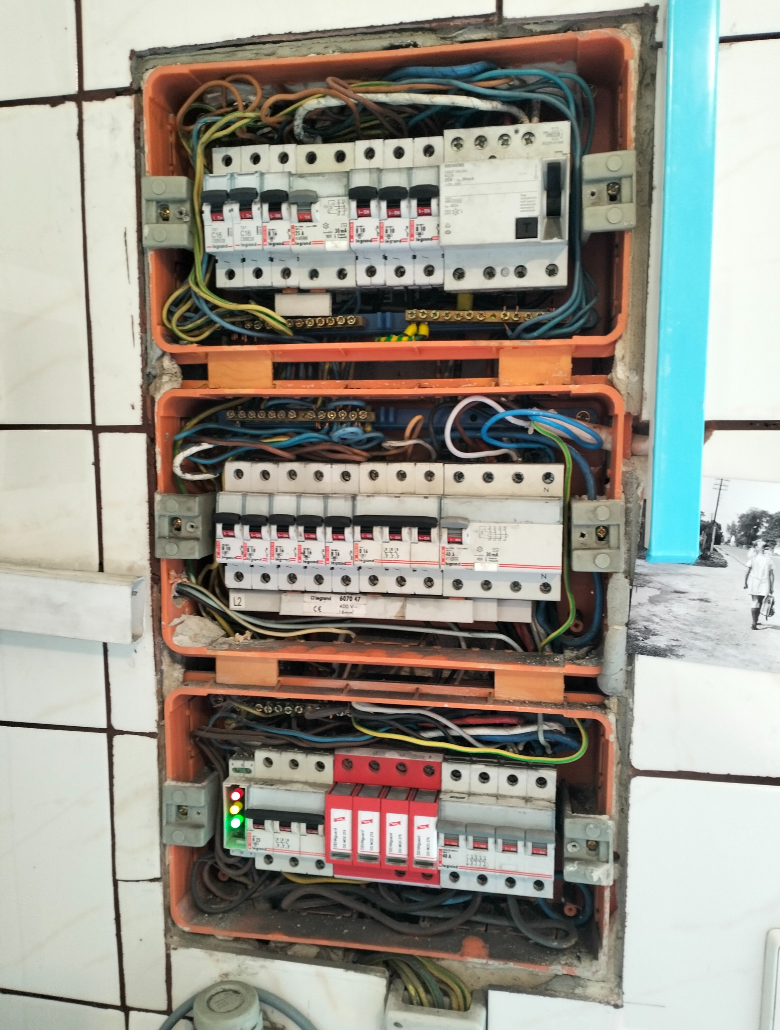 Rozdzielnia elektryczna w pomarańczowej obudowie, widoczne bezpieczniki, przewody i listwy. Instalacja zamontowana w ścianie z białymi płytkami. Widoczne elementy instalacji elektrycznej.