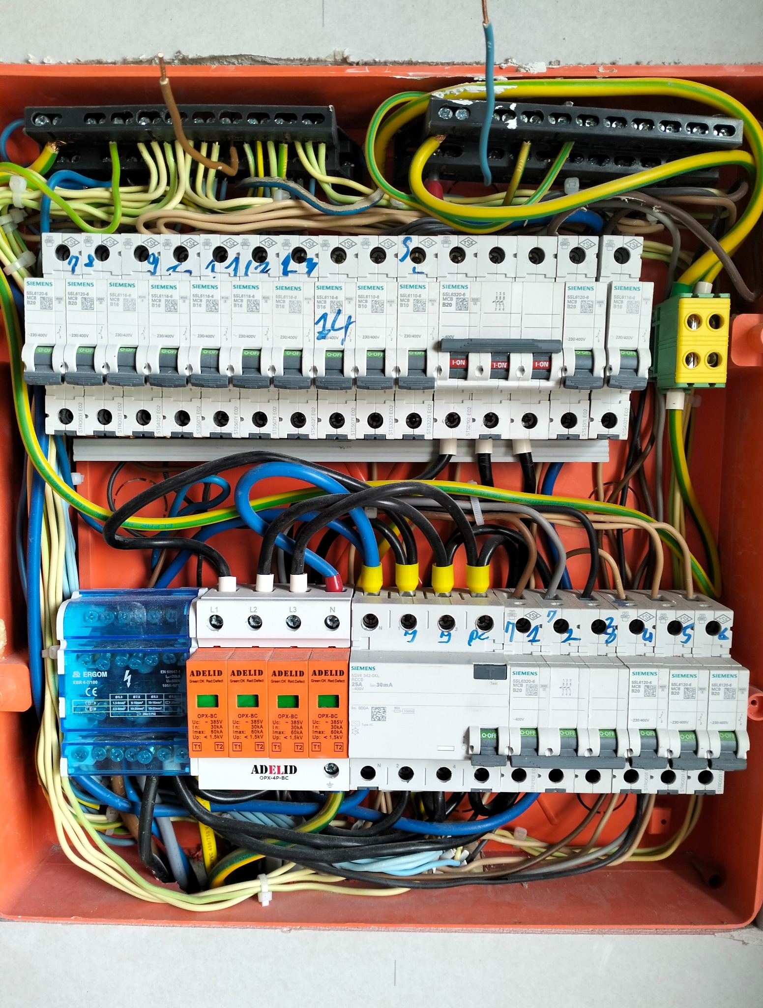 Otwarta skrzynka elektryczna z bezpiecznikami Siemens i Adelid, widoczne kable w różnych kolorach, oznaczenia na bezpiecznikach, całość w pomarańczowej obudowie.