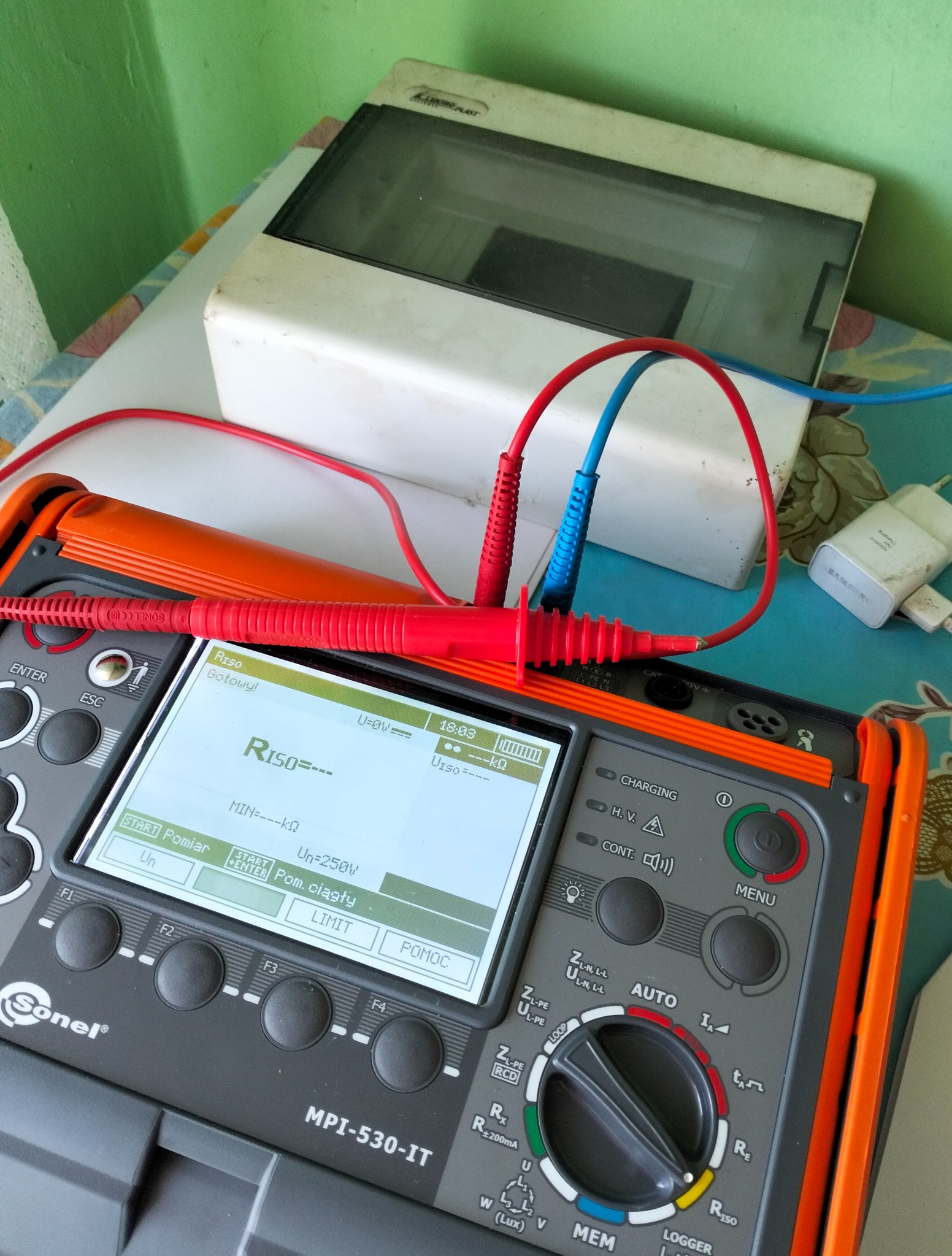 Miernik Sonel MPI-530-IT z podłączonymi przewodami pomiarowymi, wyświetlacz pokazuje wynik pomiaru rezystancji izolacji. Obok skrzynka elektryczna i ładowarka.