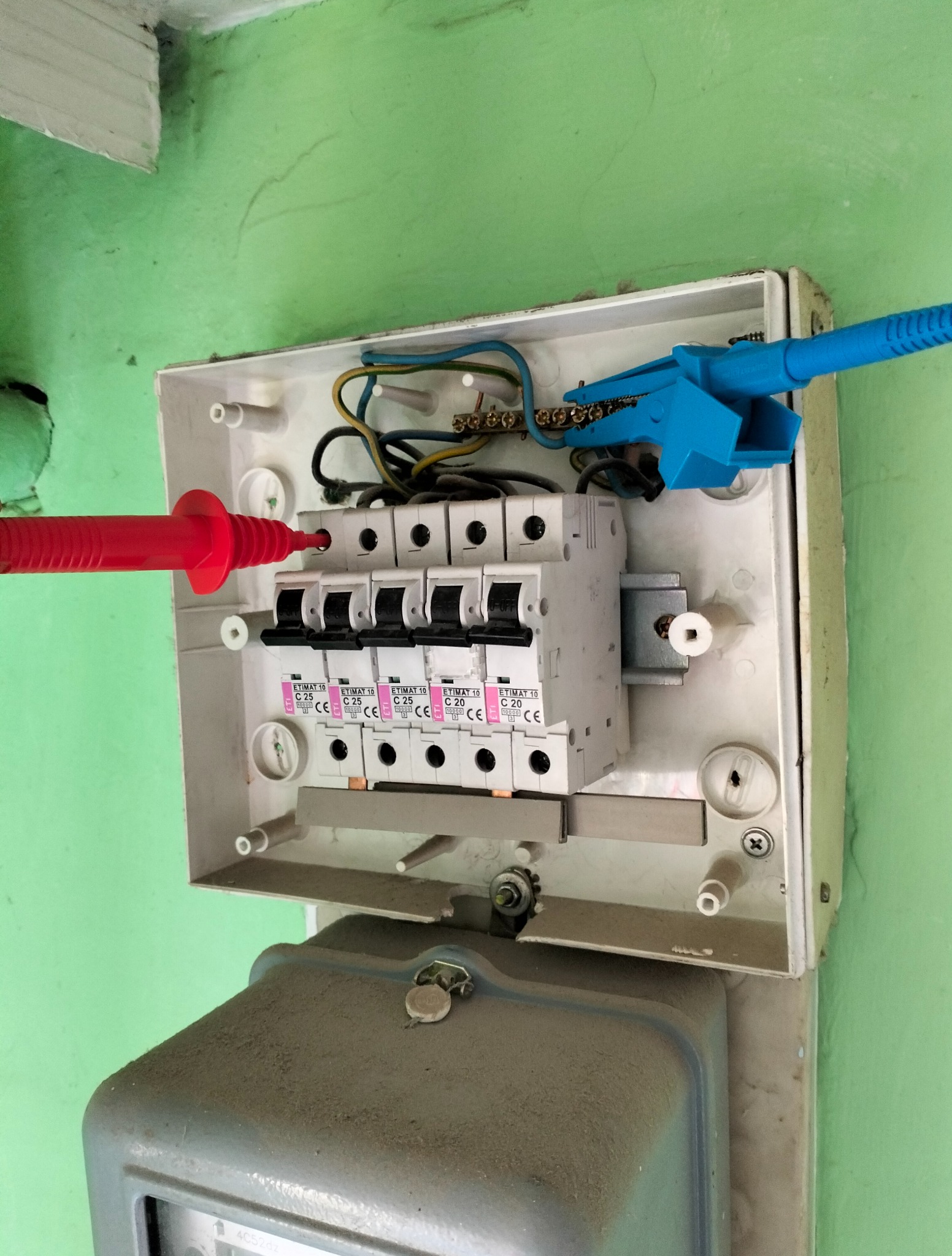 Otwarta skrzynka bezpiecznikowa z podłączonymi przewodami pomiarowymi (czerwony i niebieski) na tle zielonej ściany i szarego licznika energii.