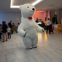 TwojeChwile360 - Duży, pluszowy biały niedźwiedź z czerwoną muszką, stoi na parkiecie w sali balowej podczas imprezy. W tle DJ i tańcząca para.