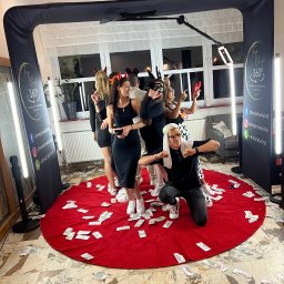 TwojeChwile360 - Wesoła grupa w przebraniach pozuje w videobudce 360° na czerwonym dywanie zasypanym rekwizytami imitującymi pieniądze. Na ściankach budki widoczne logo i dane kontaktowe.