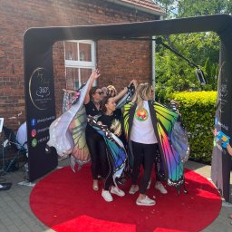 TwojeChwile360 - Videobudka 360 w Słupcy: grupa kobiet w kolorowych skrzydłach pozuje na czerwonym dywanie. W tle budynek z cegły i zieleń. Zabawa i uśmiech na evencie.