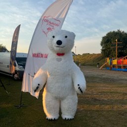 TwojeChwile360 - Duży, biały miś w kostiumie na tle flagi 'Karawaning z pasją' i placu zabaw. Maskotka z czerwoną muchą.