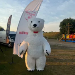 Duży, biały miś w kostiumie na tle flagi 'Karawaning z pasją' i placu zabaw. Maskotka z czerwoną muchą.